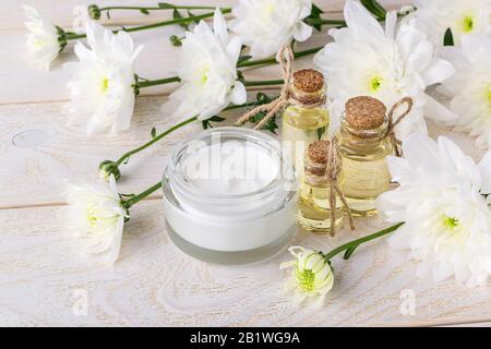 Crema viso in vaso di vetro, tre bottiglie di olio essenziale di crisantemo e crisantemi bianchi su un tavolo di legno. Bellezza, cura della pelle e cosmetologia. Foto Stock