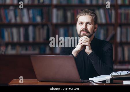 Uomo maturo bearded in collo di polo nero seduto al tavolo con libri aperti e guardando la fotocamera con sfondo sfocato. Felice maschio finito lavorare su lapt Foto Stock