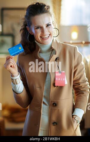 giovane donna sorridente in maglione bianco e gonna cercando cappotto beige con etichetta prezzo di vendita rosso e tenendo carta di credito blu in negozio di moda moderno. Foto Stock