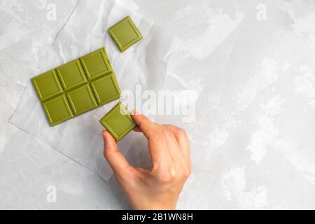 Mano della donna che tiene un pezzo di cioccolato verde del tè del matcha sullo sfondo grigio neutro. Vista dall'alto con spazio di copia Foto Stock