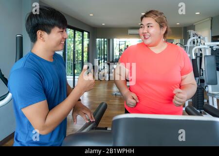 Due uomini asiatici dell'addestratore e la donna di overweight che esercitano l'addestramento sul tapis roulant nella palestra, l'addestratore che osserva felice il suo risultato ed il pollice in su durante l'allenamento. Grasso Foto Stock