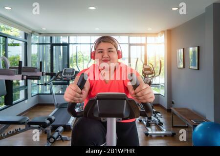 Felice Asian Overweight donna con l'allenamento degli auricolari su cyclette in palestra moderna, felice e sorridente durante l'allenamento. Donna grassa prendersi cura di salute e. Foto Stock