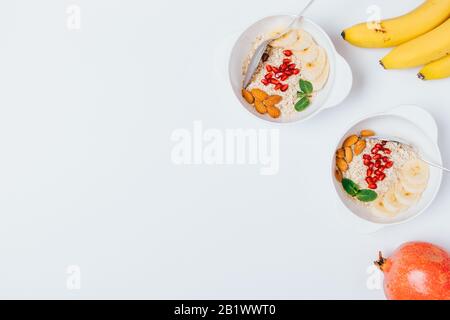 Colazione all'ingrosso di farinata d'avena con noci, semi di melograno e banane su tavola bianca, piatto con spazio copia. Foto Stock