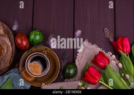 Colazione di Pasqua con caffè espresso fresco aromatico, croissant, torta brownie, uova colorate, tulipani rossi e salice. Caffè con dolci, fiori su un tavolo di legno. Vista dall'alto Foto Stock