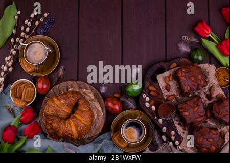 Colazione di Pasqua con caffè espresso fresco aromatico, croissant, torta brownie, uova colorate, tulipani rossi e salice. Caffè con dolci, fiori su un tavolo di legno. Vista dall'alto Foto Stock