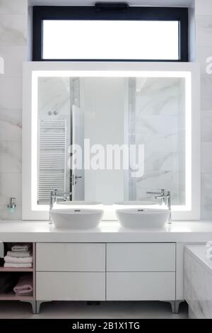 Bagno moderno bianco e pulito con due lavandini, grande specchio con cornice luminosa e finestra superiore Foto Stock