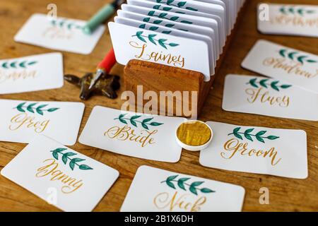 Calligraphy di nozze - schede del posto scritte a mano con i nomi. Scrittura dorata con verde fiorisce su carta bianca. Vernice oro, pennarello, porta carte di credito. Foto Stock