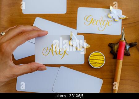 Calligraphy di nozze - le schede del posto handwritten della mano femminile con i nomi e le farfalle della carta. Scrittura dorata su white paper. Foto Stock