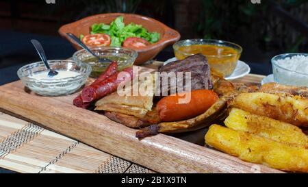 Grande vassoio di legno con carni alla griglia e salsicce accompagnate da salse, manioca fritta, un piatto di riso e pomodoro e insalata di lattuga su un tavolo nero Foto Stock