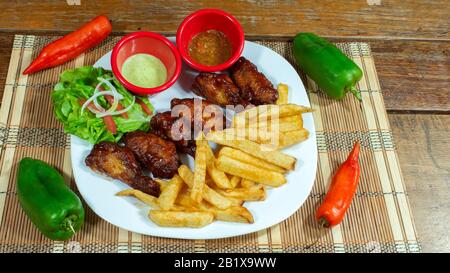 Piatto bianco con ali di pollo barbecue accompagnate da patatine fritte, salse e insalata, ornata con peperoni verdi e rossi su un tavolo di legno Foto Stock