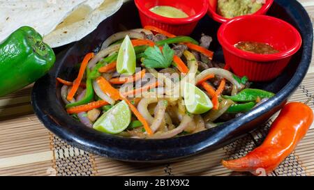 Piatto di argilla con fajitas di manzo con strisce di pepe e cipolla accompagnate da un piatto di tortillas di mais e salsa piccante, guacamole e un margarita blu su un Foto Stock