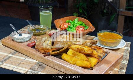 Grande vassoio di legno con carni alla griglia e salsicce accompagnate da salse, manioca fritta, un piatto di riso e pomodoro e insalata di lattuga su un tavolo nero Foto Stock