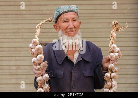 Uomo anziano uyghur con barba grigia che tiene corde di aglio in un bazar a Kashgar. L'uomo indossa un tradizionale cappello doppa. Foto Stock