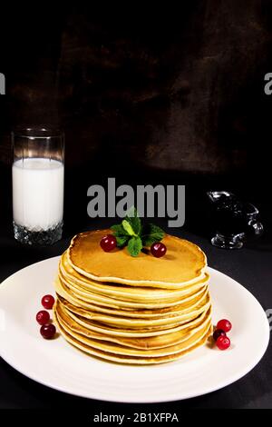 Deliziosa colazione a base di pancake appena sfornati su un piatto bianco con latte in vetro e foto Verticale di mirtilli freschi su un fondo scuro Foto Stock