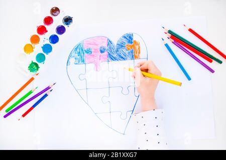 Un bambino sta disegnando cuore colorato con puzzle jigsaw su sfondo bianco come un simbolo di autismo, con matite colorate e acquerelli. Foto Stock