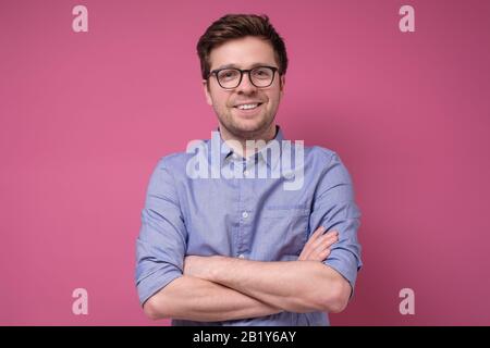 Giovane sorridente con occhiali in piedi con braccia incrociate su sfondo rosa Foto Stock