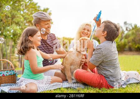 Felice famiglia facendo picnic in natura all'aperto - giovani genitori divertirsi con i bambini e il loro cane in estate ridere, giocare insieme con il sapone Foto Stock