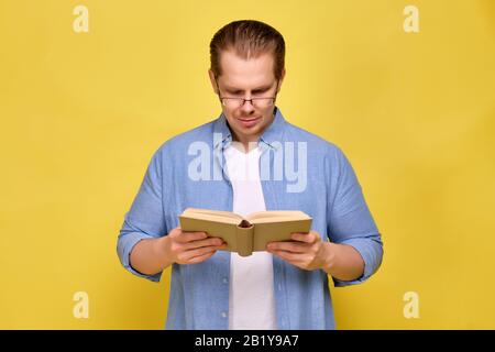 Un uomo in una camicia su uno sfondo giallo legge un libro con occhiali. Primo piano. Foto Stock