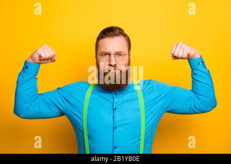 Foto di attraente divertente ragazzo sollevare seriamente braccia pugni presentare forma perfetta forte bicipite sport addicted indossare camicia blu verde sospenders Foto Stock