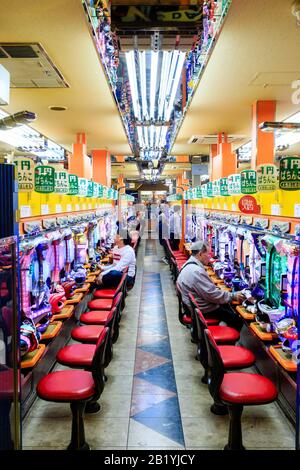 Vista interna lungo la navata di un salotto giapponese di pachinko con due file di slot machine e alcuni uomini seduti a giocare su di loro. Notte. Foto Stock