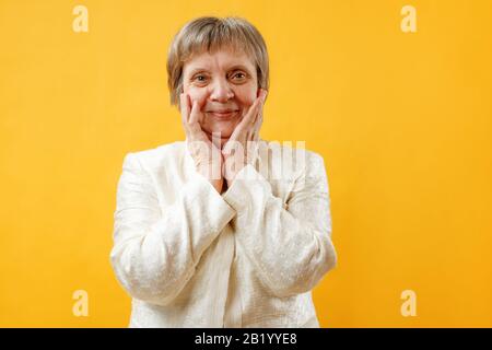 Ritratto di una donna sorridente gioiosa dai capelli grigi stupita con le palme sulle sue guance su uno sfondo giallo. Foto Stock