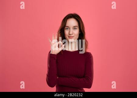 Felice giovane signora in piedi isolato su sfondo rosa scuro guarda la fotocamera che mostra il gesto di OK-simbolo. Foto Stock