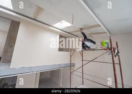 giovane operaio edile professionale che utilizza impalcature mentre intonacando su soffitto in gesso all'interno del nuovo grande appartamento moderno a due livelli Foto Stock