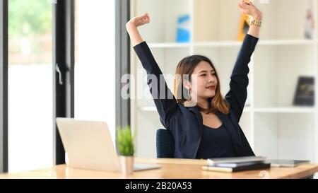 Foto di giovane bella businesswoman in abito nero formale rilassante dopo il lavoro finito al tavolo da lavoro in legno con comodo ufficio come backgr Foto Stock