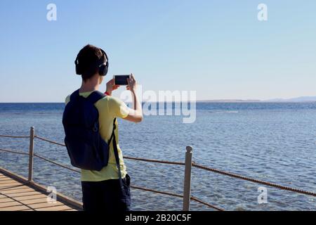 Ragazzo hippster con cuffie che scattano foto di un paesaggio incredibile su cellulare smart phone fotocamera digitale di una roccia vicino al mare, bella luce solare Foto Stock