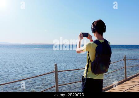 Ragazzo hippster con cuffie che scattano foto di un paesaggio incredibile su cellulare smart phone fotocamera digitale di una roccia vicino al mare, bella luce solare Foto Stock