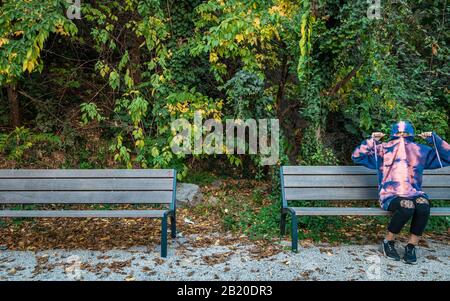 Giovane ragazza che si nasconde in una felpa con cappuccio su una panchina in un parco Foto Stock