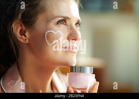 elegante e penosa donna di 40 anni in pigiama con cuore bianco crema viso su guancia tenuta vaso cosmetico rosa nella casa moderna nella soleggiata giornata invernale Foto Stock