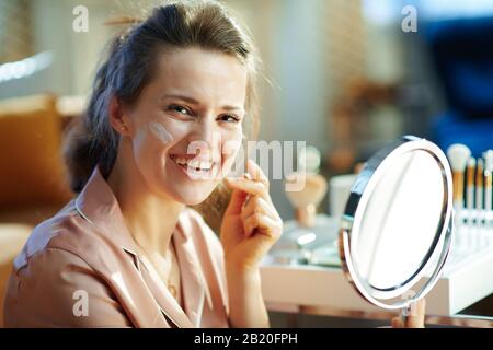 Ritratto di donna sorridente elegante in pigiama con specchio applicazione crema viso vicino al tavolo con prodotti da bagno nel moderno soggiorno in inverno da soleggiato Foto Stock