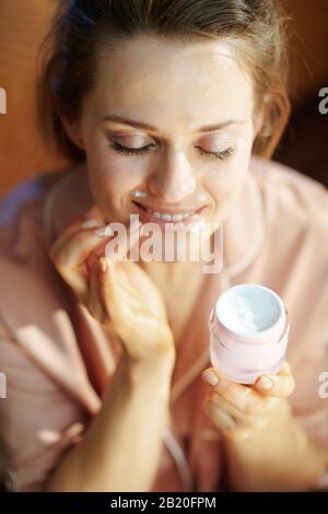 happy 40 anni casalinga in pigiama con vaso cosmetico rosa applicare labbro contorno crema in casa moderna nella giornata invernale soleggiata. Foto Stock