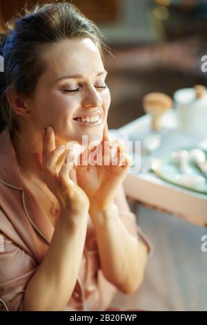 buona casalinga di mezza età in pigiami applicazione crema viso vicino al tavolo con prodotti da bagno nella casa moderna in giornata invernale soleggiata. Foto Stock