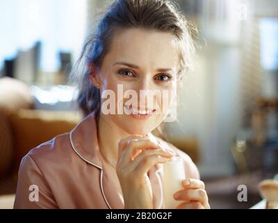 felice elegante casalinga di mezza età in pigiama con tubo cosmetico bianco a casa moderna nella soleggiata giornata invernale. Foto Stock