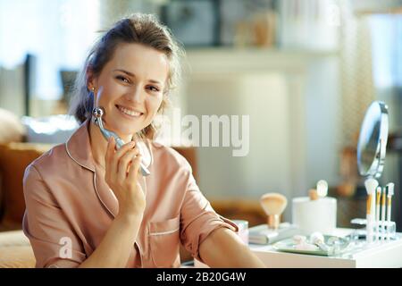 Ritratto di felice donna di 40 anni in pigiama nel moderno soggiorno nella soleggiata giornata invernale utilizzando l'ascensore facciale anti invecchiamento massaggiatore elettrico. Foto Stock