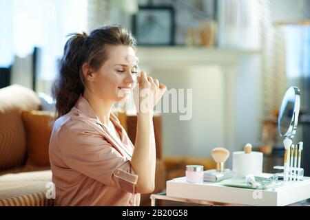 giovane donna felice in pigiami utilizzando calmante massaggiatore a rullo al quarzo vicino al tavolo con articoli da toeletta nel moderno soggiorno nella soleggiata giornata invernale. Foto Stock