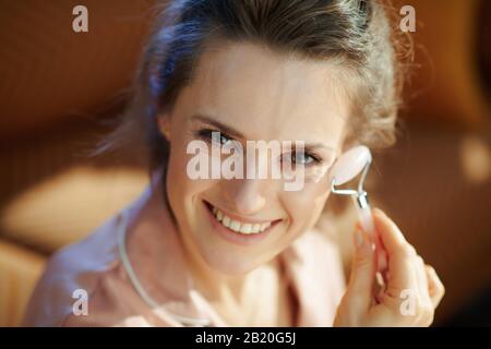 donna moderna e sorridente di 40 anni in pigiama in casa moderna nella soleggiata giornata invernale utilizzando un massaggiatore a rullo al quarzo. Foto Stock