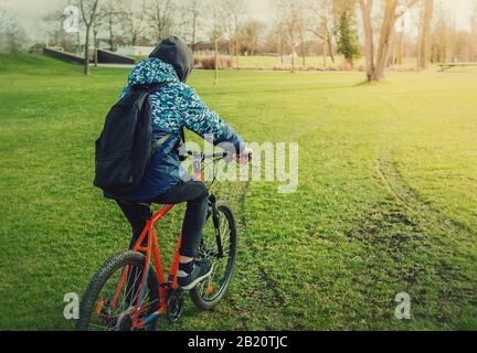 Vista posteriore di un uomo ciclista in bicicletta la mattina presto primavera nel parco. Turistico con uno zaino e felpa con cappuccio su una bicicletta. Foto Stock