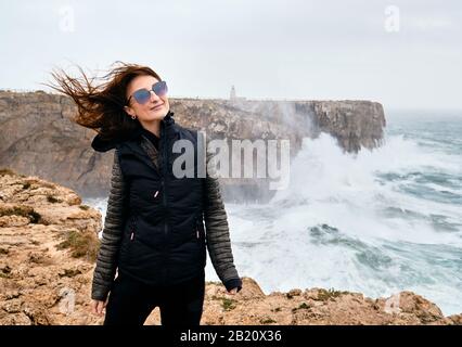 Millennial caucasica donna i suoi capelli fluttering nel vento che si posa in piedi sul bordo della scogliera, tempestoso Oceano Atlantico sullo sfondo. Fortaleza Sagres Foto Stock