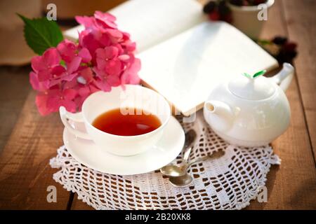 Da sopra di tazza di tè rosso e bianco ceramica vaso da tè su tavolo in legno con fiori rosa e libro aperto Foto Stock