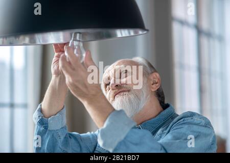 Uomo anziano concentrato che cambia una lampadina nella sua lampada Foto Stock