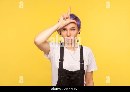 Perdente! Ritratto di ragazza ipster dispiaciuto con i capelli viola corti in tute di denim che mostrano il gesto perdente, il simbolo del dito sulla testa, abou preoccupato Foto Stock