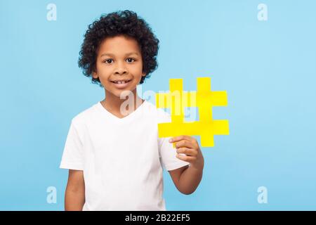 Simbolo hash. Gioioso felice ragazzo carino con capelli ricci in T-shirt tenendo grande giallo hashtag simbolo e sorridendo alla fotocamera, blogging e contenuti per bambini Foto Stock