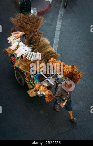 Vista dall'alto del venditore di strada che tira un carrello con scopa, spolverini e altri materiali di pulizia, vendendoli su una strada a Bangkok, Thailandia. Foto Stock