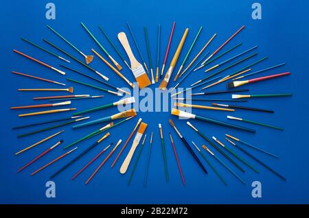 Set di pennelli per pittura d'arte di vari colori, dimensioni e tipi di setole disposti in sequenza circolare. Scatto piatto, isolato su sfondo blu Foto Stock