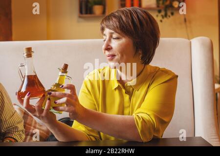 Attraente donna matura che tiene bottiglie di olio d'oliva e aceto balsamico in trattoria italiana Foto Stock