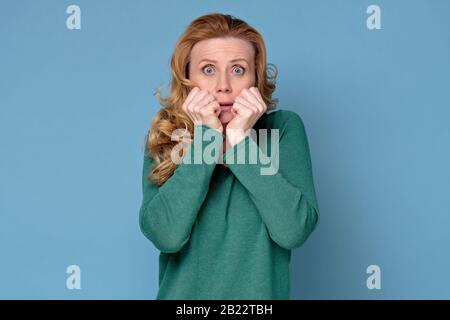 Donna caucasica matura scioccata con lunghi capelli in piedi su sfondo blu. Madre è nel panico becaue suo bambino ha fatto un errore. Studio Foto Stock