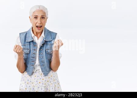 Sorpreso ed eccitato felice trionfando vecchia donna con capelli grigi, mani clench, pugno pompa che celebra la vittoria, lotteria vinta, dire sì allegramente, realizzare Foto Stock